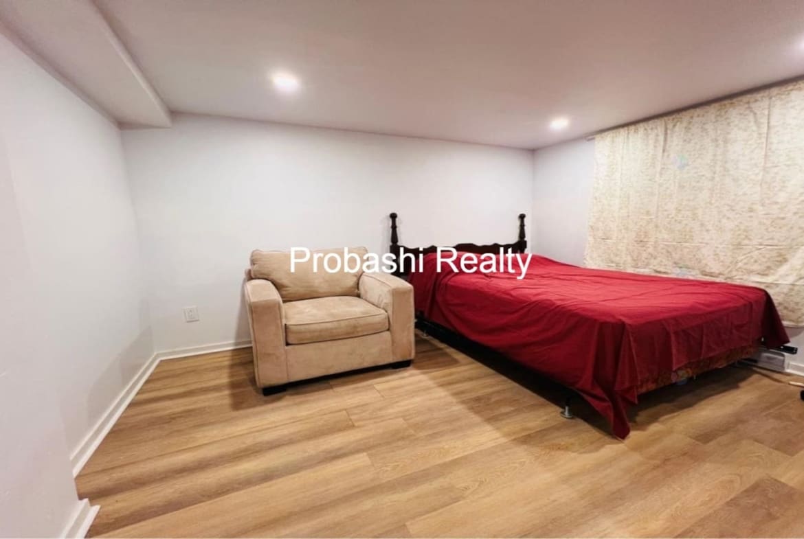Cozy bedroom with wooden flooring, beige armchair, and red bedspread.