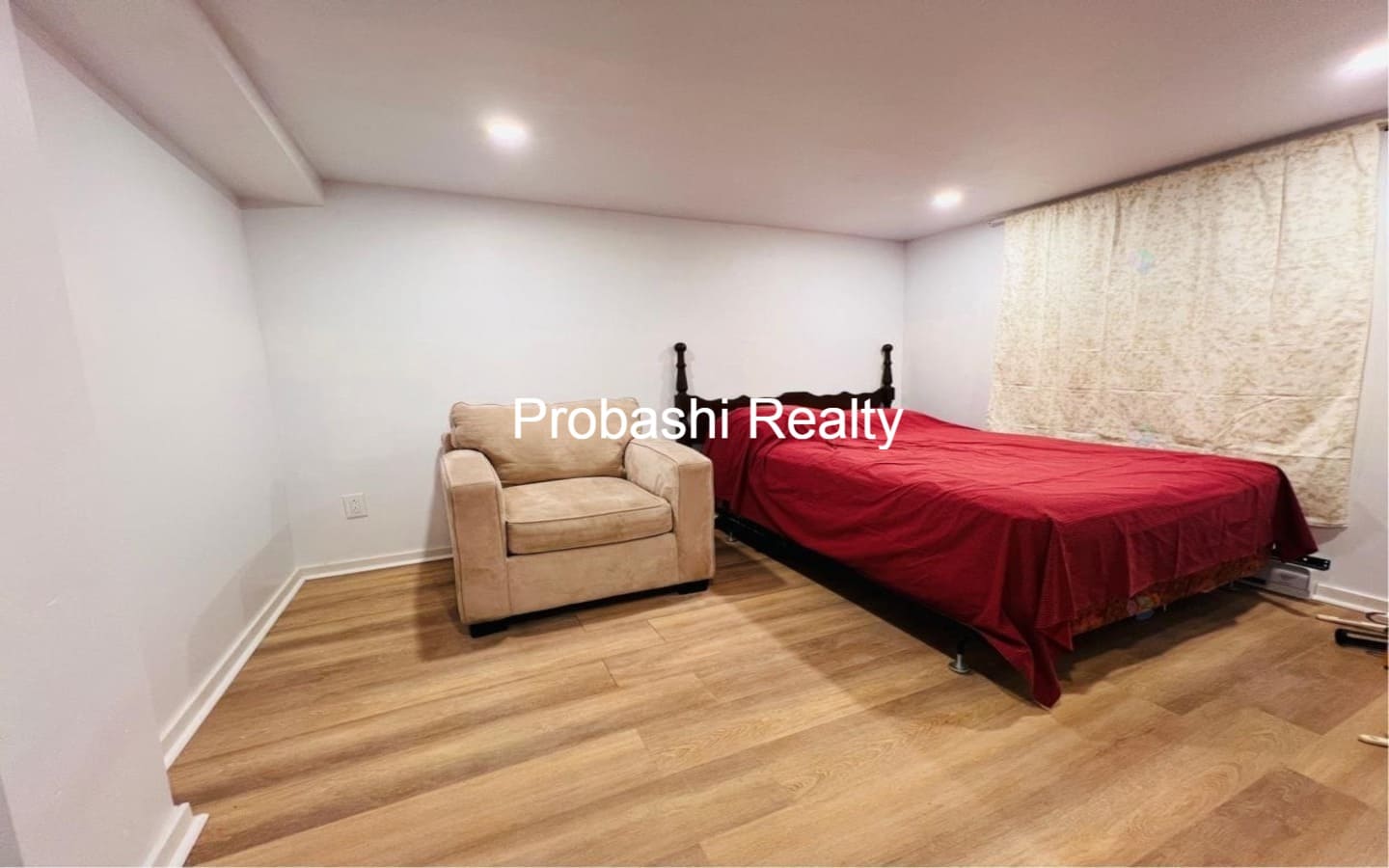Cozy bedroom with wooden flooring, beige armchair, and red bedspread.